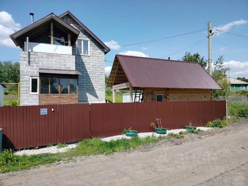 Строение выполнено в современном архитектурном стиле с использованием кирпича и дерева. Основное здание имеет два этажа, с балконом на верхнем уровне и большими окнами, обеспечивающими естественное освещение. Рядом расположена небольшая деревянная постройка с крышей из металлочерепицы, что создает контраст с основным домом. Забор из металлических панелей придает территории аккуратный вид. Цветовая гамма включает серые и коричневые оттенки, а также зеленые акценты в виде горшков с растениями, что добавляет уюта и гармонии в общий облик.