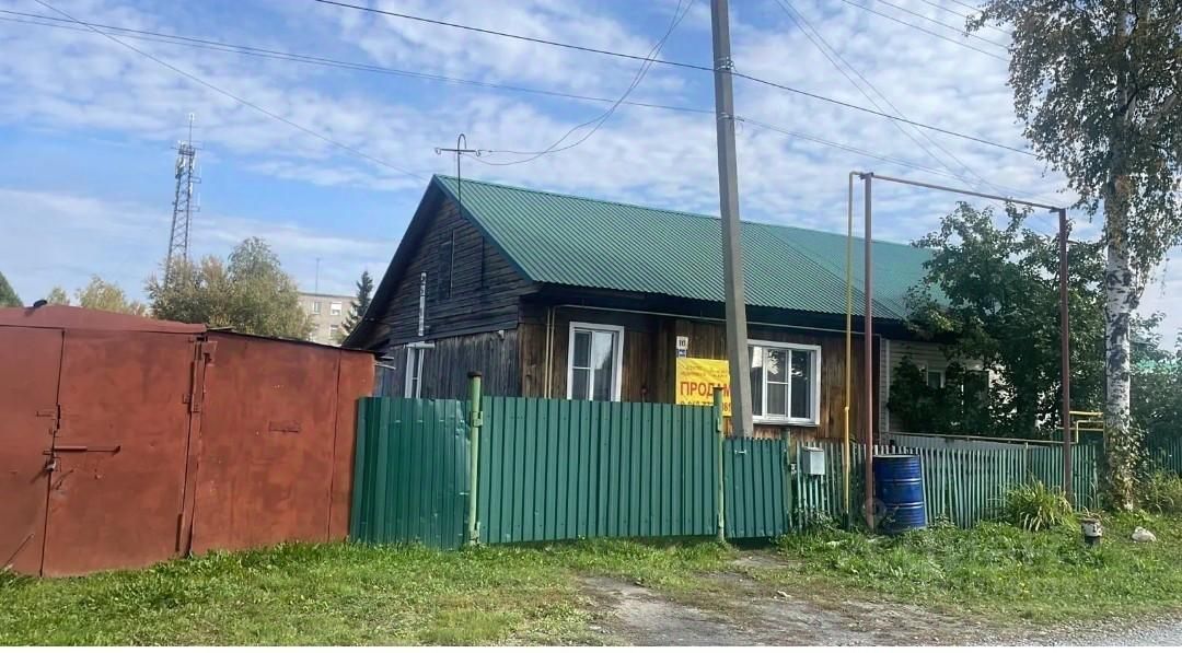 Деревянный дом в традиционном русском стиле с зеленой крышей и деревянными стенами, отделанными досками. Видны большие окна, обрамленные белыми рамами, и яркая желтая вывеска с надписью "Продается". Ограда выполнена из зеленых металлических листов, создавая уютный и закрытый участок. Слева расположен красный металлический гараж, а справа — несколько кустов и деревьев, добавляющих зелени. Общая атмосфера передает простоту и функциональность загородного жилья.