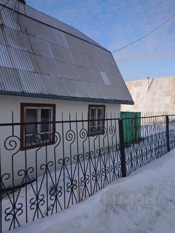 Здание выполнено в традиционном стиле с наклонной крышей, покрытой металлическими листами. Фасад окрашен в светлый цвет, окна обрамлены деревянными рамами с решетками. Видна декоративная металлическая ограда с изящными узорами, которая окружает участок. Угловая дверь зеленого цвета придает яркий акцент. Снег на земле создает контраст с темными элементами конструкции. Пространство выглядит уютным и гармоничным, отражая классические черты загородного дома.