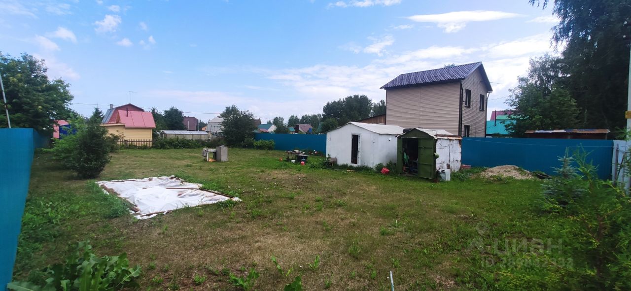 На фотографии представлен участок земли с ухоженной травой и несколькими постройками. В центре виден белый сарай с зеленой дверью, а рядом расположены стол и стулья, создающие пространство для отдыха. Слева можно заметить накрытый белым материалом участок, возможно, для хранения. Вдалеке виден двухэтажный дом с коричневой крышей и светлыми стенами. Участок окружен высоким забором, что придает уединение. Зеленые кустарники и деревья добавляют естественности и создают уютную атмосферу. Общая палитра включает зеленые, белые и коричневые оттенки.