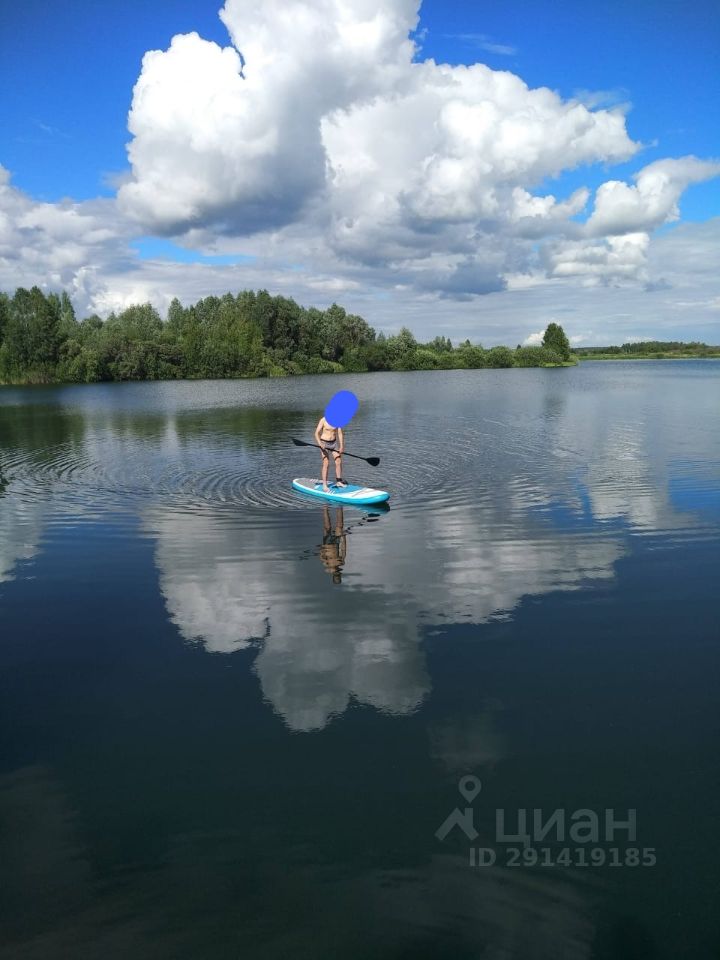 На фотографии видно человека, стоящего на доске для серфинга с веслом на спокойной воде. Вода отражает облака и зелень вокруг, создавая живописный пейзаж. Человек одет в купальные шорты, а доска имеет яркий голубой цвет. Облака на небе крупные и пушистые, создавая контраст с ярким небом. Вдали виднеются деревья, которые добавляют природный элемент к сцене. Общее ощущение спокойствия и отдыха на воде.
