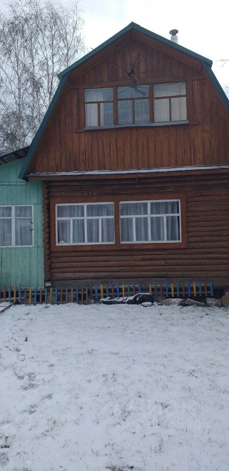 Архитектурный стиль дома представлен в виде сочетания деревянной и окрашенной в светлый цвет фасадной отделки. Структурные особенности включают двускатную крышу с высокими фронтонами и большие окна, обеспечивающие естественное освещение. Видимая часть здания демонстрирует деревянные панели и элементы из бруса, что придает ему традиционный деревенский стиль. Основные характеристики включают широкие окна с белыми занавесками, создающими уютную атмосферу. Цветовая гамма включает натуральные древесные оттенки и светлые тона, что гармонично сочетается с окружающей средой.