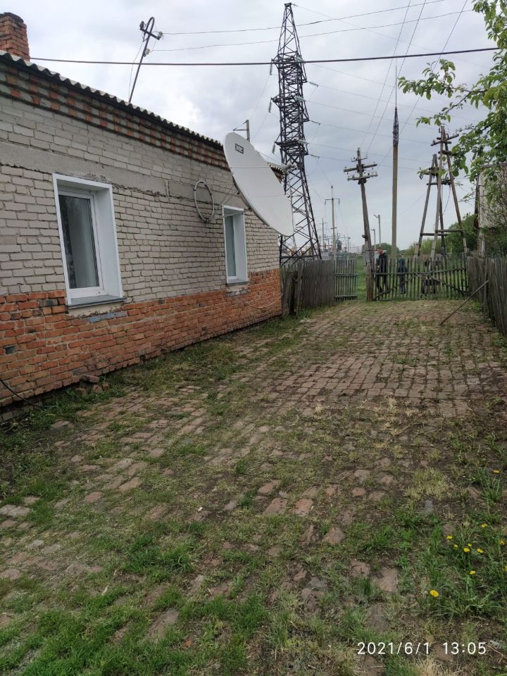 Стена дома выполнена из кирпича с частично оштукатуренной поверхностью. Видны два окна, обрамленные белыми рамами. На стене расположена спутниковая антенна, что указывает на наличие современных коммуникаций. Пол во дворе выложен из кирпичей, образующих ровную поверхность, с участками травы и поросли. Общий вид пространства создает ощущение уюта и простоты, характерных для загородного жилья. Отсутствие крупных объектов мебели или декора подчеркивает открытость и функциональность данного участка.