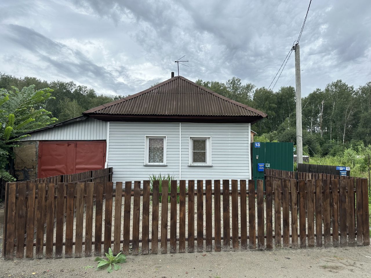 Дом в стиле дачи с односкатной крышей, покрытой металлочерепицей. Стены обшиты светло-голубым сайдингом, окна имеют простые рамки и занавески. Входная дверь красного цвета, рядом с ней расположены ворота. Забор из деревянных досок создает уютное ограждение. Участок вокруг дома пустой, с небольшими растениями. Основные цвета — голубой, красный и коричневый. Простота и функциональность дизайна подчеркивают характер загородного жилья, создавая атмосферу уюта и спокойствия.