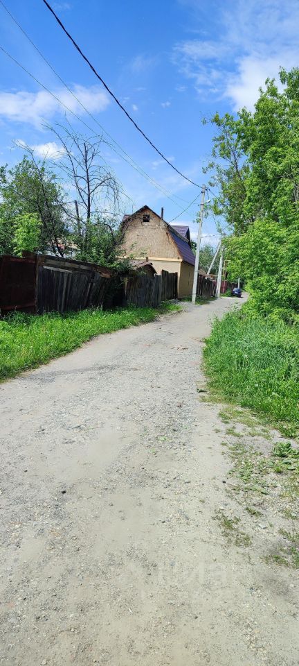 На фотографии видно узкую грунтовую дорогу, окруженную зелеными насаждениями и деревьями. С одной стороны дороги расположены деревянные заборы, а с другой стороны виден дом с крышей, покрытой черепицей. Стены дома выполнены из кирпича, что придает ему прочный вид. Дорога слегка извивается, создавая ощущение уединения. Вдали можно заметить столбы электропередач, что указывает на наличие коммуникаций в этом районе. Цвета в основном натуральные: коричневые, зеленые и серые, что гармонирует с окружающей природой.