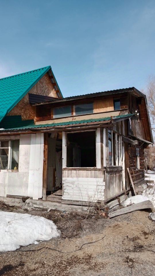 Здание выполнено в традиционном стиле с элементами деревянного строительства. Крыша с зелёной черепицей придаёт характерный вид. Видны открытые пространства, где отсутствуют стены, что создаёт ощущение недостроенности. Внутреннее пространство, вероятно, предназначено для жилых нужд, однако состояние требует ремонта. Использованы деревянные и бетонные материалы, что подчеркивает rustic стиль. Цвета преобладают натуральные: коричневый от дерева и белый от штукатурки. Финишные детали, такие как оконные рамы и двери, частично отсутствуют, что добавляет элемент незавершенности.