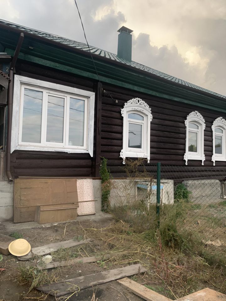 Деревянный дом в традиционном русском стиле с характерной отделкой. Стены выполнены из тёмного бруса, что придаёт ему уютный вид. Окна оформлены белыми рамами с изящными резными наличниками, что подчеркивает архитектурные особенности. Крыша с зелёной черепицей, имеющая небольшие скаты, добавляет шарма. Видна часть фундамента, выполненная из серого бетона. Вокруг дома растут кустарники и трава, создавая естественную атмосферу. Общая цветовая гамма включает тёмные и светлые оттенки, гармонично сочетающиеся друг с другом.