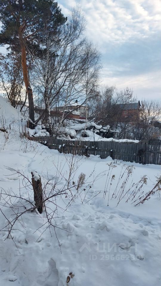 На фотографии виден участок с покрытым снегом ландшафтом. В центре расположены обнаженные ветки и кустарники, обрамленные снежным покровом. На заднем плане виднеется деревянный забор, который ограждает территорию. За забором просматриваются строения, выполненные в традиционном стиле, с крышей, наклоненной к одной стороне. Цвета в изображении преимущественно холодные, с преобладанием белого и серого, создающими зимнюю атмосферу. Структурные элементы, такие как деревья и забор, добавляют глубину и текстуру в общую композицию.