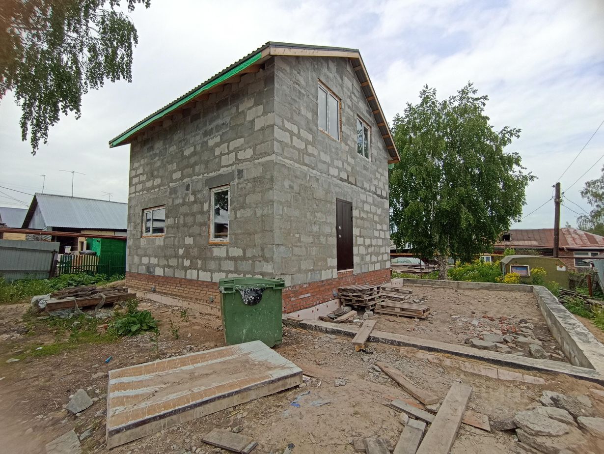 Стена здания выполнена из бетонных блоков, с частично открытой кирпичной отделкой внизу. На фасаде видны окна с деревянными рамами, а также входная дверь. Крыша с зелёным покрытием добавляет контраста к серым тонам стен. Вокруг здания разбросаны строительные материалы, такие как деревянные поддоны и плиты, что указывает на незавершённый этап строительства. Площадка перед домом не выровнена, на ней видны следы работы, что создаёт ощущение активного строительства. Общая композиция демонстрирует современный подход к архитектуре с элементами функциональности.