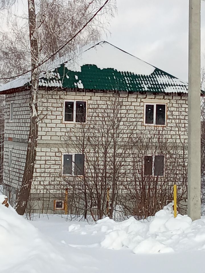 Двухэтажное здание выполнено в простом архитектурном стиле с кирпичной отделкой. Крыша с зеленой черепицей, частично покрытой снегом, создает контраст с белыми стенами. Окна прямоугольной формы, установлены в симметричном порядке, придают фасаду аккуратный вид. На первом этаже видны элементы, указывающие на наличие подвала. Стены из светлого кирпича, что подчеркивает простоту и функциональность конструкции. Вокруг здания расположены кустарники и деревья, что добавляет уюта, несмотря на зимний пейзаж.