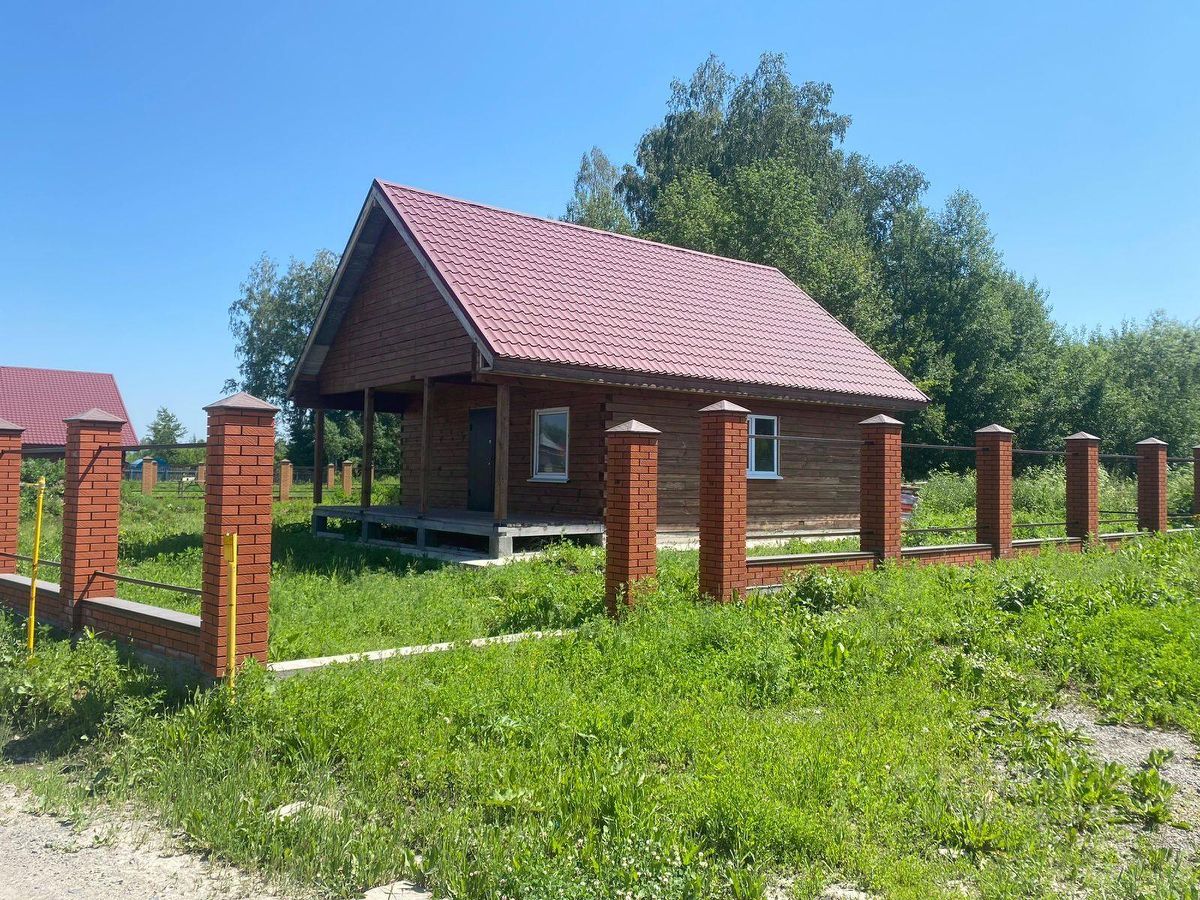 Деревянный дом в загородном стиле с красной крышей из металлочерепицы. Архитектурный дизайн включает в себя выступающие элементы и открытый крыльцо, поддерживаемое столбами. Стены отделаны натуральным деревом, придающим уютный вид. Окна имеют простой, но элегантный дизайн, обеспечивая естественное освещение. Забор выполнен из красного кирпича, создавая четкую границу участка. Цветовая палитра включает теплые деревянные оттенки и яркий красный цвет крыши, что делает дом привлекательным и гармоничным с окружающей природой.