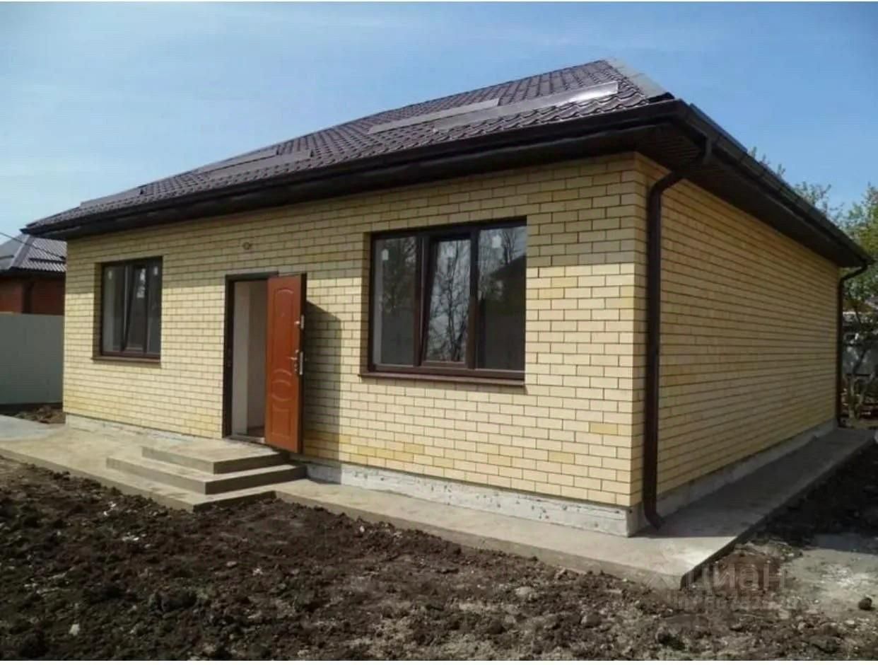 Дом выполнен в современном архитектурном стиле с простыми линиями и практичным дизайном. Фасад облицован светло-желтым кирпичом, что придает ему уютный вид. Структурные особенности включают симметричное расположение окон и входной двери, а также наклонную крышу с темной черепицей. Входная группа оформлена массивной деревянной дверью с металлической ручкой. Окна большие, с темной рамой, что создает контраст с кирпичной кладкой. Отделка фасада и крыши выполнена в спокойных, нейтральных тонах, что делает дом визуально привлекательным и современным.