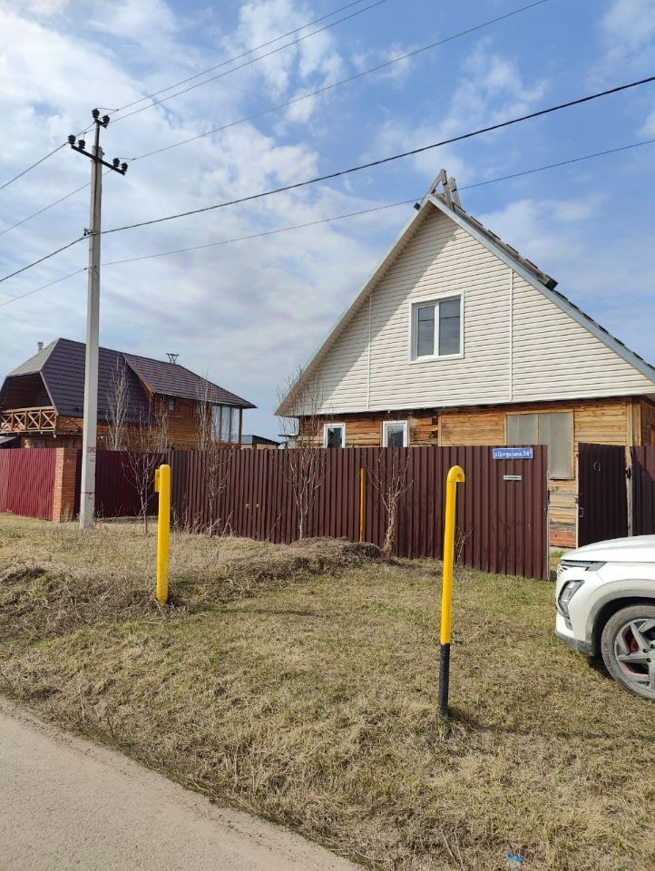 Деревянный дом в загородном стиле с двускатной крышей и светлыми стенами. Фасад дома отделан светлым сайдингом, окна имеют простую форму. Объект окружён забором из металлических панелей, окрашенных в тёмно-красный цвет. На переднем плане видны жёлтые столбы, возможно, для освещения. Просторный участок с небольшой растительностью, что создаёт ощущение уюта и приватности. Структурные элементы, такие как крыша и окна, подчеркивают функциональность и стиль загородного жилья.