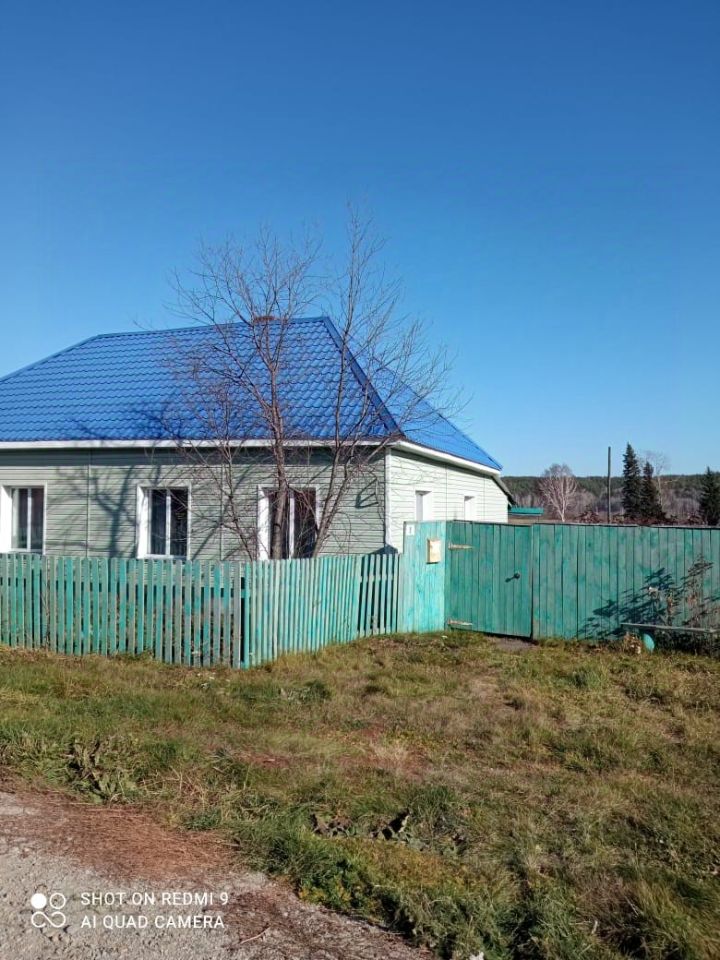 Дом в стиле дачи с яркой синей крышей и светло-зеленым фасадом. Структура одноэтажная, с симметричными окнами, обрамленными белыми рамами. Забор выполнен из деревянных досок, окрашенных в яркий зеленый цвет, создавая контраст с фасадом. Видно ухоженное пространство перед домом с небольшим газоном. Окна имеют простую, но привлекательную форму, что подчеркивает уютный характер здания. Деревянные элементы забора и фасада добавляют естественности и гармонии в общий вид.