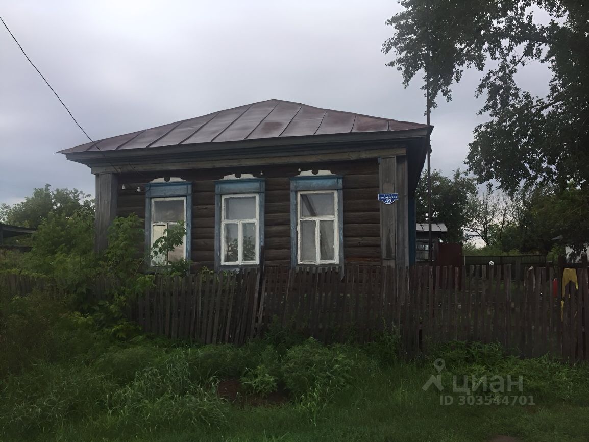 Деревянный дом в русском стиле с характерной архитектурой. На фасаде видны большие окна с белыми рамами и синими наличниками, что придает зданию яркий акцент. Крыша имеет двускатную форму и выполнена из металлического материала. Стены дома состоят из бревен, что подчеркивает традиционный стиль постройки. Огороженная территория с деревянным забором создает уютную атмосферу. Зелень вокруг здания добавляет живописности, а также создает ощущение уединения и спокойствия.