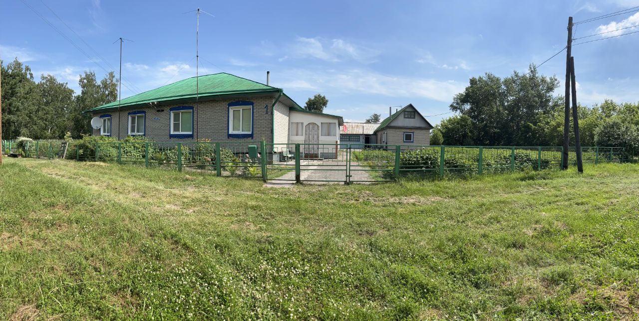 Дом выполнен в традиционном стиле с зеленой крышей и кирпичной отделкой. Окна обрамлены синими рамами, что придает зданию яркий акцент. Структура включает в себя одноэтажное здание с отдельным входом и небольшим крыльцом. Перед домом находится ухоженный участок с зеленым забором, а также дорожка, ведущая к входу. Внешние стены имеют светлый оттенок, что гармонирует с крышей. Общая атмосфера создает ощущение уюта и спокойствия, а также подчеркивает простоту и функциональность дизайна.