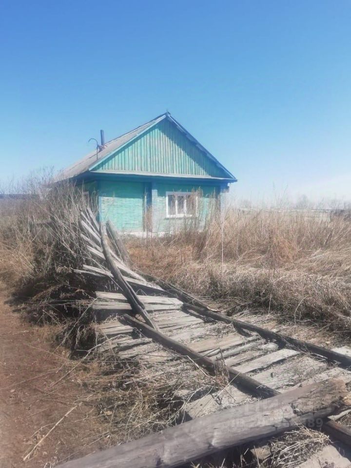 Деревянный дом в стиле русской деревенской архитектуры с зеленой окраской фасада. Крыша двускатная, покрыта шифером. Окна с белыми рамами, придающими уютный вид. Перед домом виден неухоженный участок с заросшими травами и старой деревянной дорожкой, частично разрушенной. Деревянные элементы конструкции, такие как балки и доски, демонстрируют естественные текстуры и цвета древесины. Общая атмосфера пространства создает ощущение уединения и простоты, характерное для сельской местности.