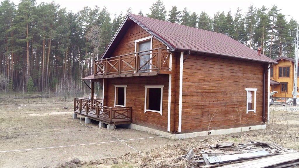 Деревянный дом в загородном стиле с наклонной крышей и балконом. Фасад отделан натуральным деревом, что придаёт ему уютный вид. Окна имеют простые белые рамы, а балкон оформлен перилами из дерева. Структура здания включает в себя открытые террасы, что создаёт дополнительное пространство для отдыха на свежем воздухе. Цвета в основном тёплые и натуральные, с преобладанием коричневых и белых оттенков. Видимые элементы архитектуры подчеркивают гармонию с природой, создавая атмосферу уединения и спокойствия.