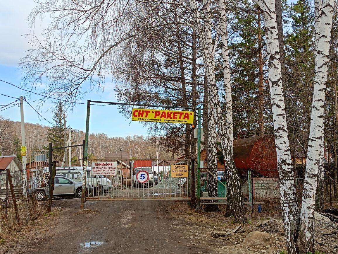На фотографии виден вход в дачный кооператив с названием "СНТ 'Пакета'". Ворота обрамлены металлической конструкцией, на которой размещен яркий рекламный баннер. Перед воротами расположены автомобили, а также видны элементы ограждения. Дорога, ведущая к кооперативу, покрыта гравием и имеет неровности. С обеих сторон дороги растут березы, создавая естественную тень. Вдалеке виднеются дачные домики, выполненные из дерева, с простыми крышами. Общая атмосфера места отражает спокойствие и уединение загородной жизни.