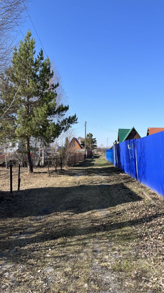 На фотографии изображена узкая дорожка, окруженная зелеными деревьями и забором. С одной стороны виден ярко-синий забор, а с другой — деревянные заборы, которые ограждают участки. На заднем плане расположены дома с крышей зеленого и коричневого цвета, выполненные в традиционном стиле. Дорожка имеет грунтовое покрытие, на котором видны следы от прохода. Вокруг растет трава, местами видны сухие листья. Пространство выглядит тихим и уединенным, создавая ощущение загородного отдыха.