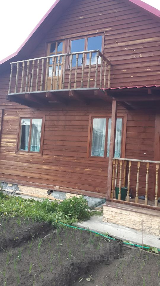 Деревянный дом в загородном стиле с балконом и крышей красного цвета. Фасад отделан деревянными панелями, создающими теплую атмосферу. Окна с рамами из дерева, оформлены шторами. На первом этаже расположены большие окна, позволяющие много света. Балкон на втором этаже поддерживается деревянными перилами, что добавляет уют. Внизу виден каменный цоколь, который контрастирует с основным материалом. Перед домом расположена небольшая клумба с зелеными растениями, создающая гармонию с природой.