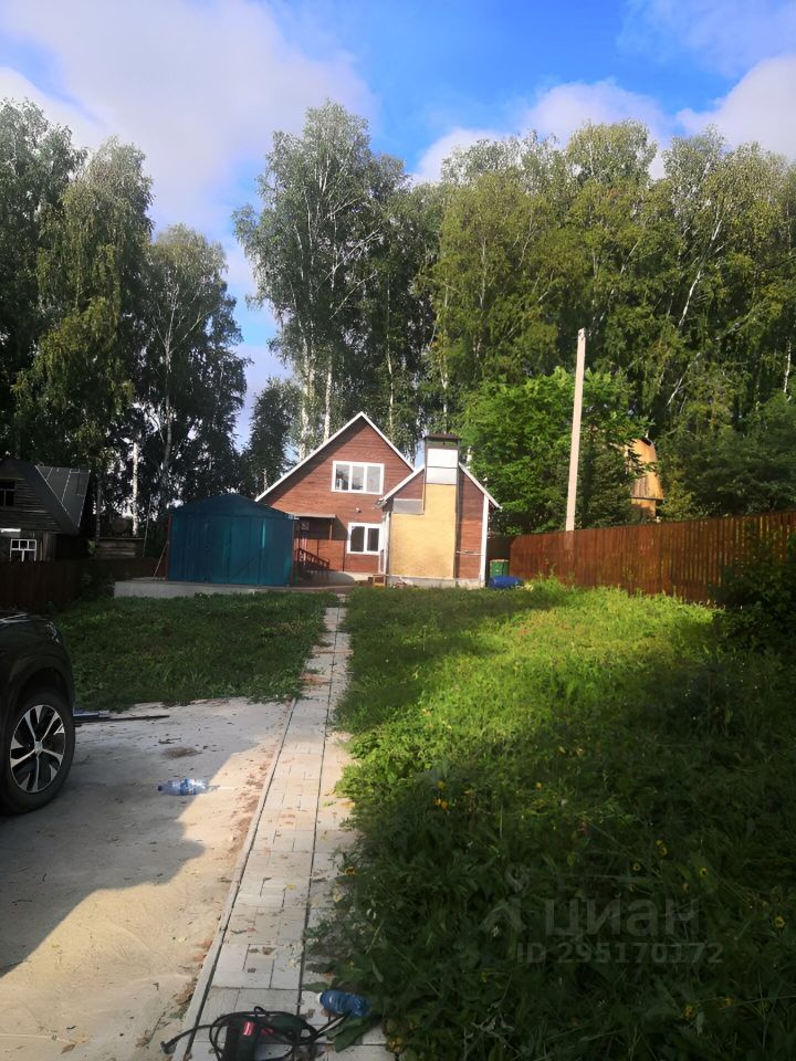 На фотографии представлен загородный дом в современном архитектурном стиле с элементами деревянной отделки. Структура включает два этажа, с мансардной крышей и большими окнами, обеспечивающими естественное освещение. Видно, что дом имеет террасу, которая выходит на зеленую лужайку. Основные материалы — дерево и кирпич, в теплых оттенках коричневого и желтого. Внешние отделки аккуратные, с акцентом на гармонию с природным окружением. Просторная территория вокруг дома, с плиточным покрытием и зелеными насаждениями, создаёт уютную атмосферу для отдыха.