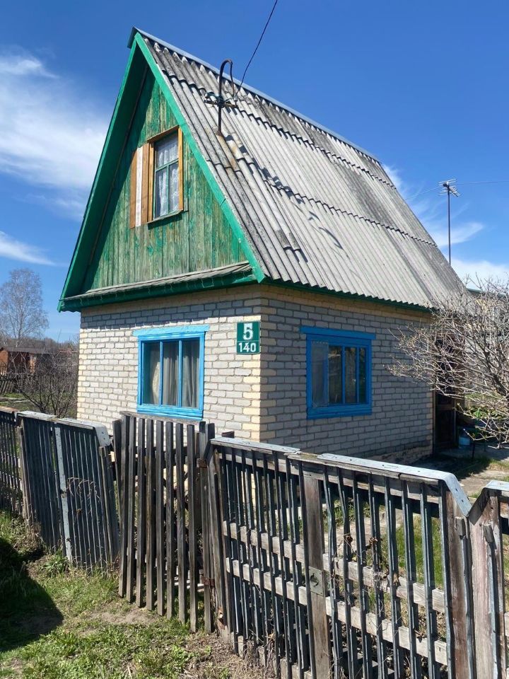 Дом в стиле дачи с двускатной крышей и кирпичной отделкой. Яркие голубые оконные рамы контрастируют с серыми кирпичами. Крыша покрыта металлическим материалом, на ней видны следы износа. Окна имеют деревянные ставни, что придаёт уютный вид. Фасад оформлен простым, но привлекательным дизайном, подчеркивающим функциональность. Забор из деревянных досок обрамляет участок, создавая ощущение уединения. Общая цветовая палитра включает серые, голубые и зеленые оттенки, что делает дом гармоничным и приятным для глаз.