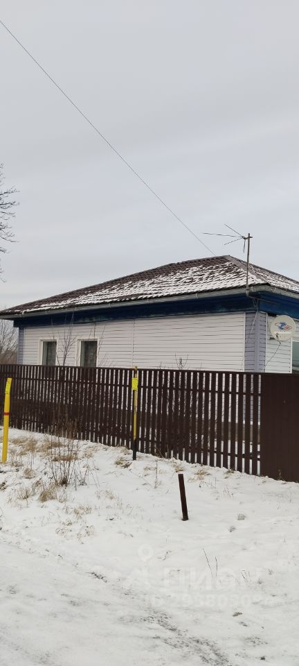 Дом в традиционном стиле с односкатной крышей и деревянной отделкой. Фасад окрашен в светлые тона, что создает уютный вид. Окна имеют простую форму, обрамлены белыми рамами, что подчеркивает аккуратность дизайна. Забор из деревянных панелей обеспечивает приватность и гармонично сочетается с общим стилем. Крыша покрыта черепицей, что добавляет характер. Пространство вокруг дома выглядит ухоженным, с небольшими участками снега, что подчеркивает зимнюю атмосферу. Доступность коммуникаций, таких как антенна, свидетельствует о современном удобстве.