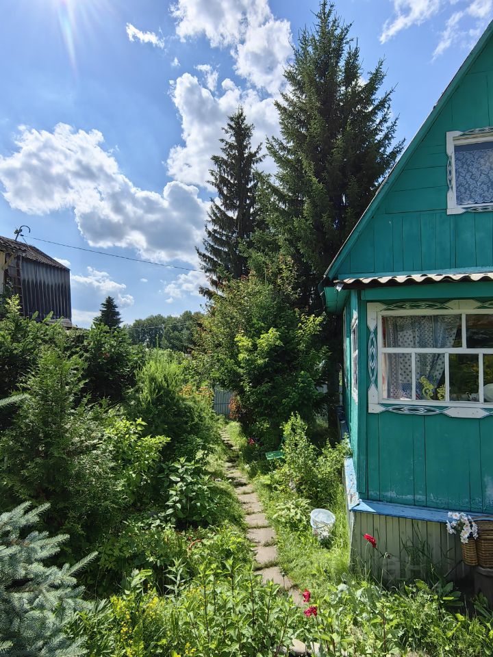 На фотографии представлен дом в традиционном русском стиле с ярко-зеленым фасадом и белыми рамами окон. Основные архитектурные элементы включают резные наличники и крытую веранду. Структурные особенности включают деревянные стены и двускатную крышу. Видна дорожка из камней, ведущая к входу, окруженная пышной растительностью. Основные характеристики пространства включают уютный и приглашающий вид, а также разнообразие зеленых растений и цветов, создающих атмосферу уединения. Цвета в интерьере гармонируют с природой, а отделка из дерева подчеркивает экологичность и традиционность.