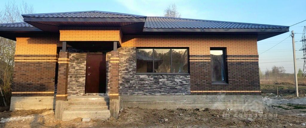 Фасад дома выполнен в современном архитектурном стиле с использованием кирпичной кладки и каменной отделки. Основные структурные элементы включают симметричное расположение окон и входной двери, а также выступающий козырек над входом. Видны большие окна, обеспечивающие естественное освещение, и контрастные горизонтальные полосы, добавляющие визуальную динамику. Используемые материалы — кирпич теплых тонов и декоративный камень, создающие гармоничный облик. Цветовая палитра включает коричневые и серые оттенки, что придает дому современный и уютный вид.