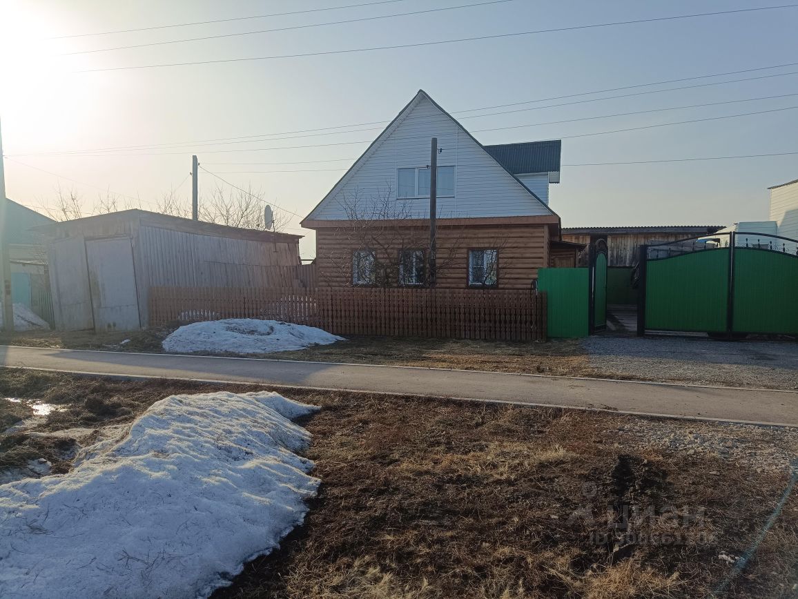 Деревянный дом с мансардной крышей и большими окнами, оформленный в традиционном стиле. Фасад выполнен из светлого дерева, что придаёт уютный вид. Перед домом расположен забор из деревянных панелей с зелёными воротами, создающий ощущение уединения. Внутреннее пространство, вероятно, открытое и светлое, с возможностью естественного освещения. На переднем плане видны остатки снега и сухая трава, что указывает на переходный сезон. Общая атмосфера вызывает ощущение спокойствия и простоты загородной жизни.