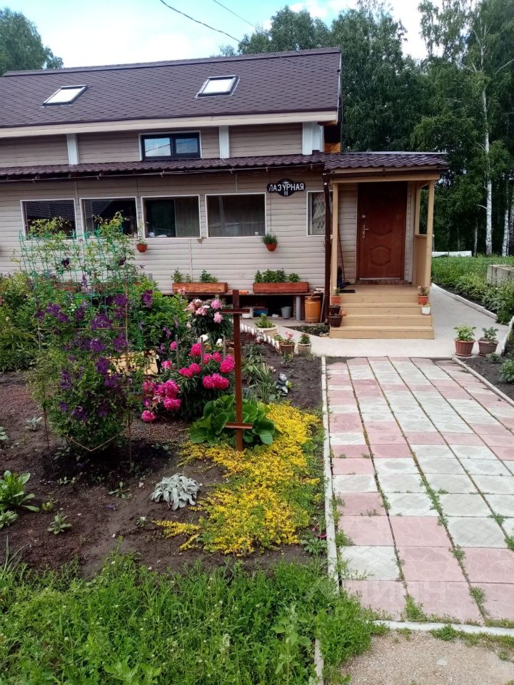 Здание выполнено в современном загородном стиле с деревянной отделкой и двускатной крышей. Входная группа оформлена лестницей и крытой верандой, что создает уютную атмосферу. Перед домом расположены цветочные клумбы с яркими растениями, включая пионы и фиалки, а также аккуратная дорожка из плитки, выполненная в светлых тонах. Использованы натуральные материалы, такие как дерево и камень, что подчеркивает гармонию с природой. Цветовая палитра включает теплые бежевые и коричневые оттенки, создавая ощущение уюта и комфорта.
