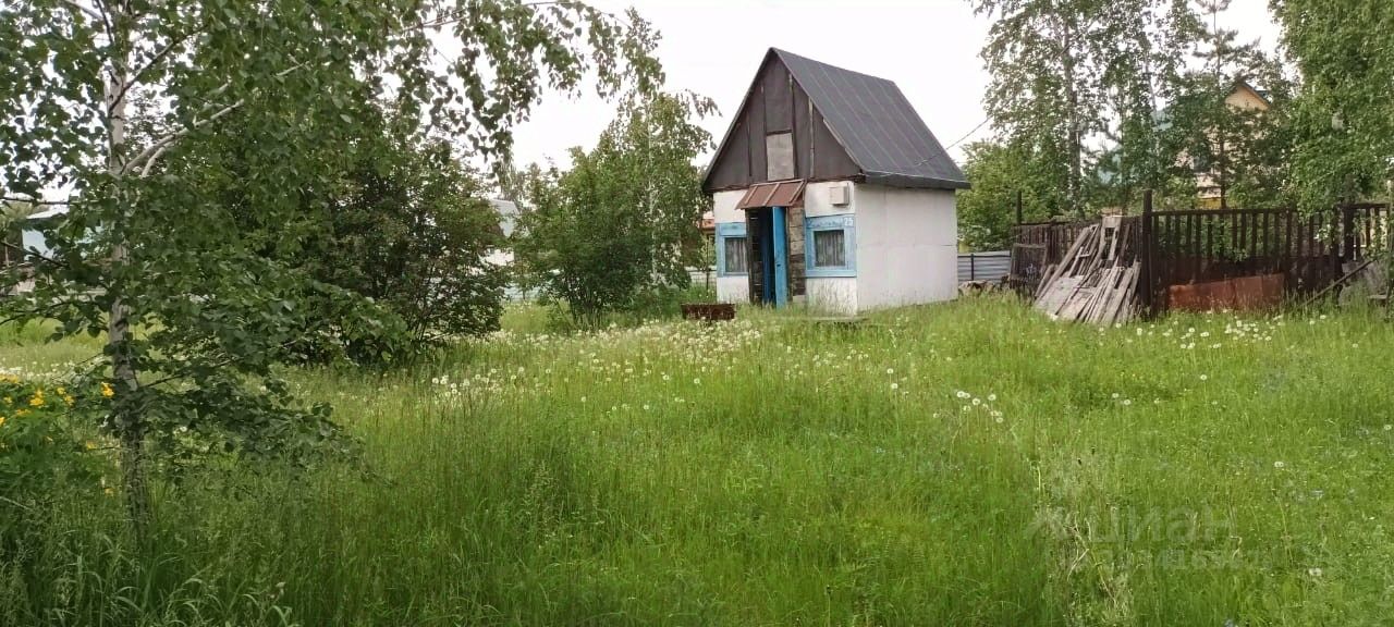 Небольшой дом с двускатной крышей, выполненный в простом архитектурном стиле. Фасад окрашен в белый цвет с синими оконными рамами и дверью. На крыше видны металлические элементы, что придаёт дому современный вид. Дверь имеет навес, а рядом расположены окна с простыми шторами. Вокруг дома зелёная трава и кустарники, создающие уютную атмосферу. Внутренние элементы не видны, однако дом выглядит компактным и функциональным, подходящим для дачного отдыха. Вдали виднеется забор и другие строения, но они не являются центральными в этом изображении.