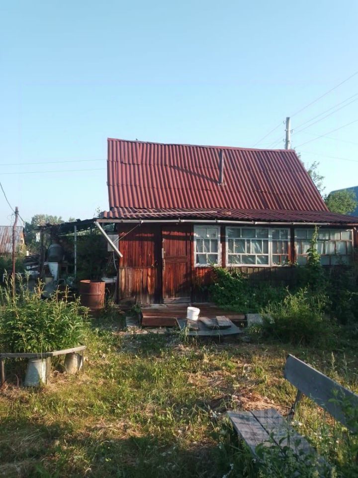 Деревянный дом с красной металлической крышей и большими окнами, оформленный в традиционном стиле. На переднем плане видна деревянная терраса, ведущая к входной двери. Стены здания отделаны темным деревом, что придает ему уютный вид. Вокруг дома растут высокие травы и кустарники, создавая естественный ландшафт. На террасе расположены простые деревянные скамейки, а также ведро и другие предметы, указывающие на использование пространства. Общая атмосфера — спокойствие и связь с природой.