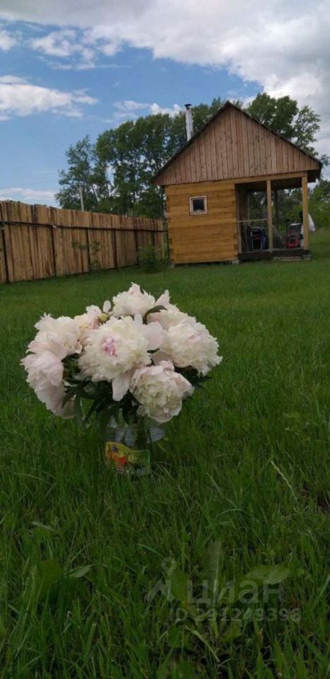 Деревянный дом в загородном стиле с простыми архитектурными линиями и крышей с небольшим скатом. Стены отделаны светлым деревом, создавая уютный вид. Перед домом расположена небольшая терраса с дверью, ведущей внутрь. На переднем плане виден букет пионов в стеклянной вазе, добавляющий яркие акценты к зелёной траве. Окна дома небольшие, что придаёт постройке традиционный вид. В целом, пространство выглядит гармонично и привлекательно для загородного отдыха.