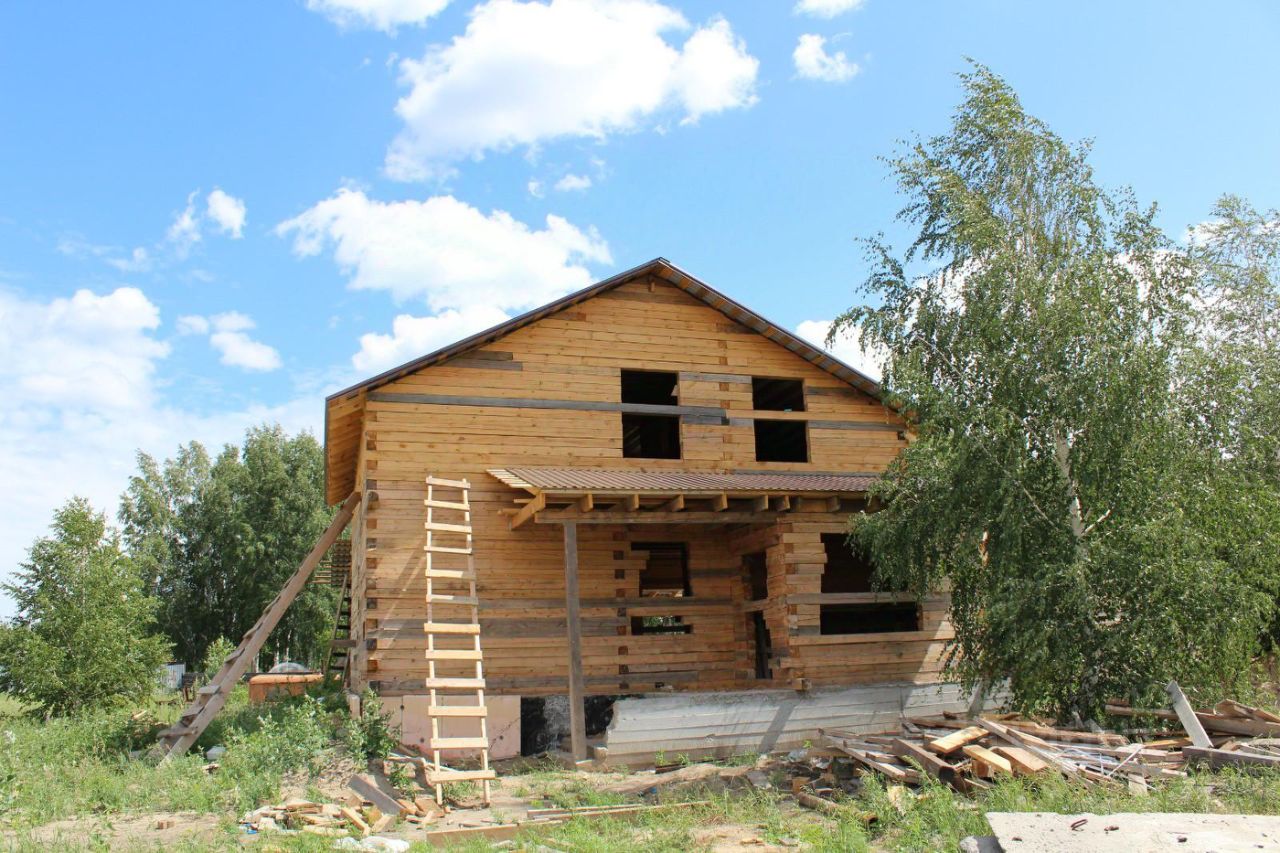 Деревянный дом в стиле загородной архитектуры с неокрашенной древесиной. Стены частично обшиты, открывая структуру бруса. На фасаде видны большие окна, которые придают светлую атмосферу. Крыша с небольшим наклоном, выполненная из металлочерепицы. Входная зона имеет навес, поддерживаемый колоннами. Лестница ведет к верхнему уровню, что указывает на многоуровневую планировку. Окружающая территория содержит строительные материалы и инструменты, что свидетельствует о процессе завершения строительства. Цвета натуральные, с преобладанием светлых древесных оттенков.