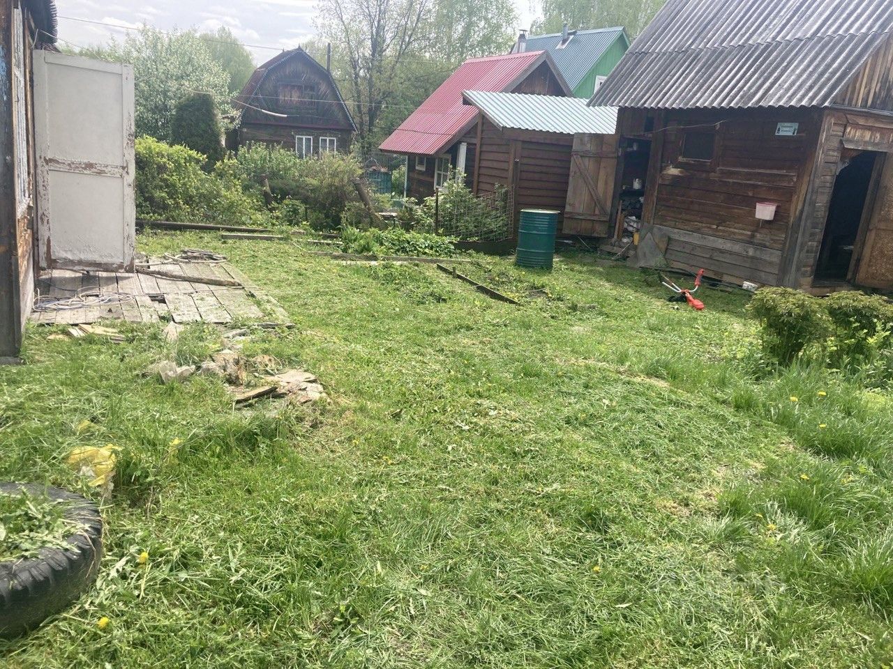 На фотографии виден участок земли с ухоженной травой и несколькими постройками. Стены деревянных домов имеют характерный rustic стиль, некоторые из них обшиты досками. Один из объектов имеет открытые двери и частично виден внутренний интерьер. Пол в этом месте состоит из деревянных досок, некоторые из которых повреждены. В углу расположен металлический контейнер и видны садовые инструменты. Цветовая палитра включает натуральные оттенки дерева и зелень травы, создавая атмосферу уюта и простоты. Пространство выглядит функциональным и подходит для дачного отдыха.