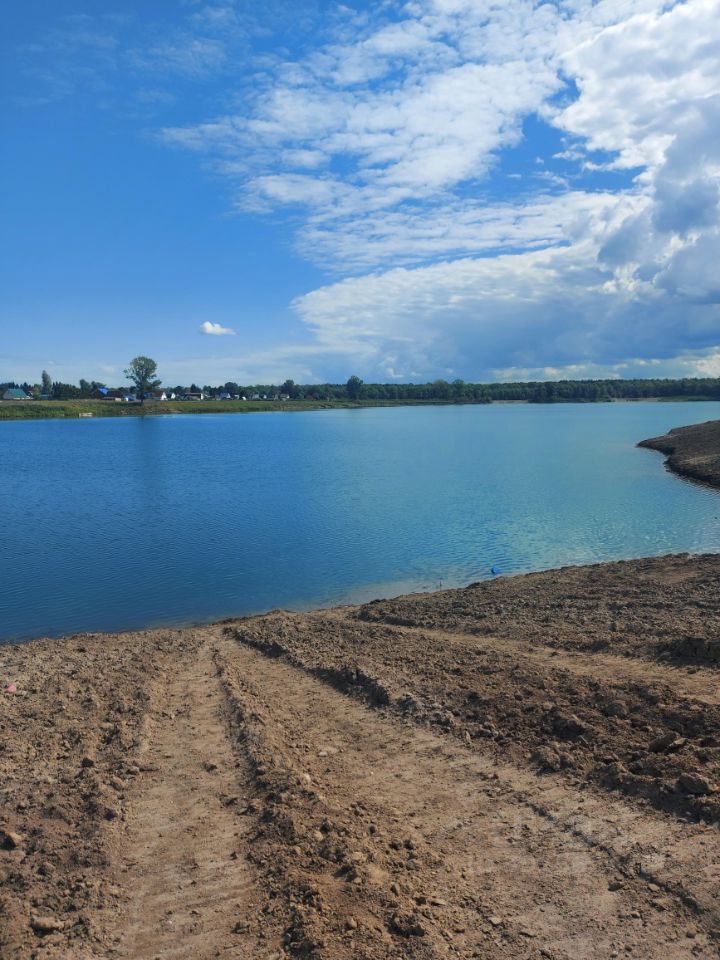 На фотографии виднеется водоем с спокойной поверхностью воды, отражающей небо. Береговая линия обрамлена рыхлой землей, с видимыми следами от техники, создающими дорожку к воде. Вдали просматриваются деревья и строения, создавая ощущение уединенности. Цвета в изображении яркие: насыщенный синий воды контрастирует с коричневыми и серыми тонами земли. Облака на небе добавляют динамичности, создавая мягкий свет. Пространство выглядит открытым и просторным, что делает его привлекательным для отдыха и активного времяпрепровождения.