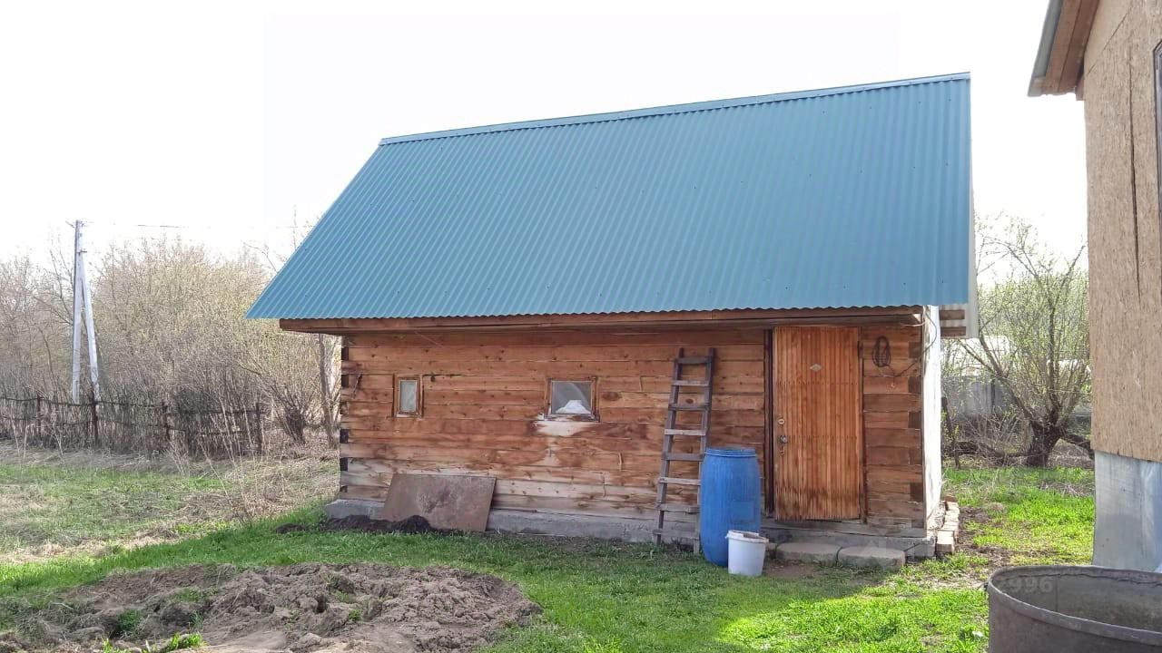 Деревянная постройка с наклонной крышей, покрытой металлическим листом синего цвета. Стены имеют натуральный деревянный отделочный материал, что придаёт дому уютный вид. На фасаде расположены небольшие окна, обеспечивающие естественное освещение. Входная дверь выполнена из дерева с простой отделкой. Слева от двери находится лестница, ведущая к крыше. Вокруг постройки видны элементы ландшафта, такие как земля и трава, но они не являются центральными в этом описании. Общая атмосфера дома создаёт ощущение уединения и простоты.