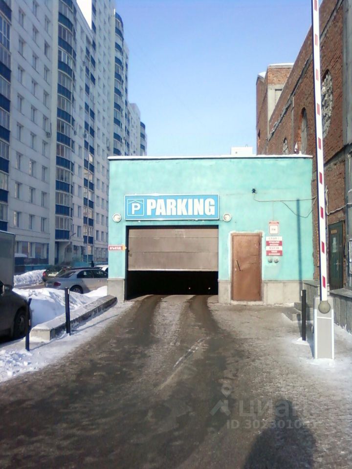 Видна парковочная зона с большими воротами, окрашенными в светло-голубой цвет. На стене расположена яркая вывеска с надписью 'PARKING'. Вход в парковку оформлен простыми архитектурными элементами, без излишеств. Справа от ворот находится дверь, выполненная из дерева, с простой ручкой. Дорога перед входом покрыта снегом и асфальтом, что придаёт пространству функциональный вид. Слева видны автомобили, припаркованные вдоль дороги. Стены здания имеют текстуру, что придаёт характер и контраст с гладкой поверхностью ворот.