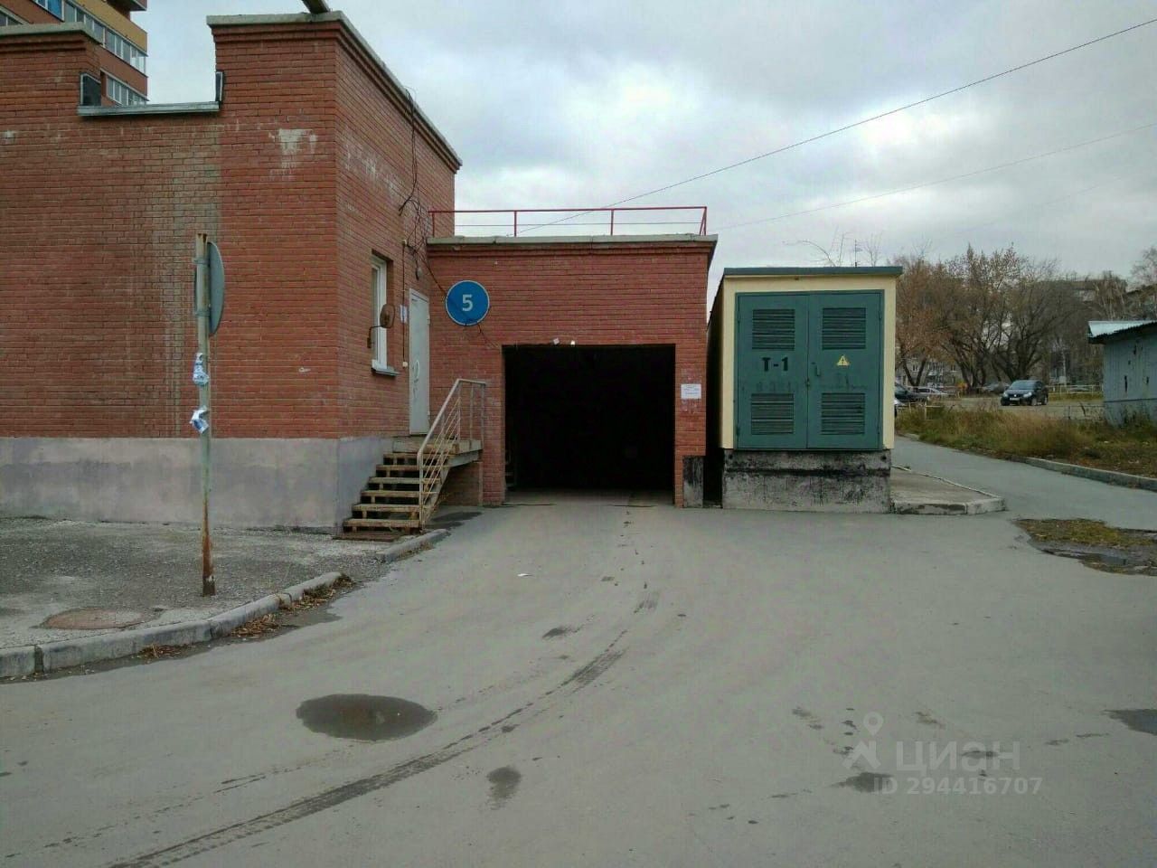Здание выполнено в кирпичном стиле с элементами промышленной архитектуры. Видны структурные особенности, такие как лестница, ведущая к двери, и открытый гараж с черным проемом. Основное пространство представляет собой вход в гараж, который имеет широкий проем и прямые линии. Цветовая гамма включает красный кирпич и серый бетон, создавая контраст. Финишные детали включают металлические элементы, такие как двери и электрический щит, что подчеркивает функциональность и практичность данного пространства.