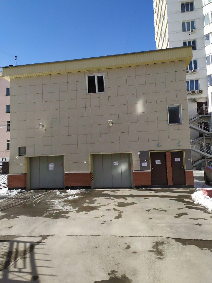 Здание выполнено в современном архитектурном стиле с гладкой фасадной плиткой светлого оттенка. На фасаде расположены три гаражные двери и несколько окон, обеспечивающих естественное освещение. Структурные элементы включают кирпичный цоколь и простые линии, создающие минималистичный вид. В верхней части здания виден светлый карниз. Пол перед зданием выложен бетонной плиткой, на которой видны следы от снега. Основные цвета – светло-желтый и бежевый, что придает зданию современный и аккуратный вид.