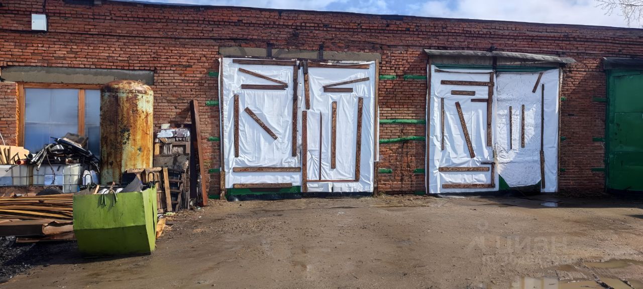 Стена из красного кирпича с тремя большими дверями, частично закрытыми белыми пленками и деревянными брусками. В центре виден металлический резервуар с ржавчиной, а также груды строительных материалов и обрезков. На переднем плане расположены зеленые контейнеры, придающие пространству индустриальный вид. Цвета в основном теплые и землистые, с преобладанием красного, зеленого и коричневого. Общая атмосфера пространства создает ощущение заброшенности и ожидания ремонта.