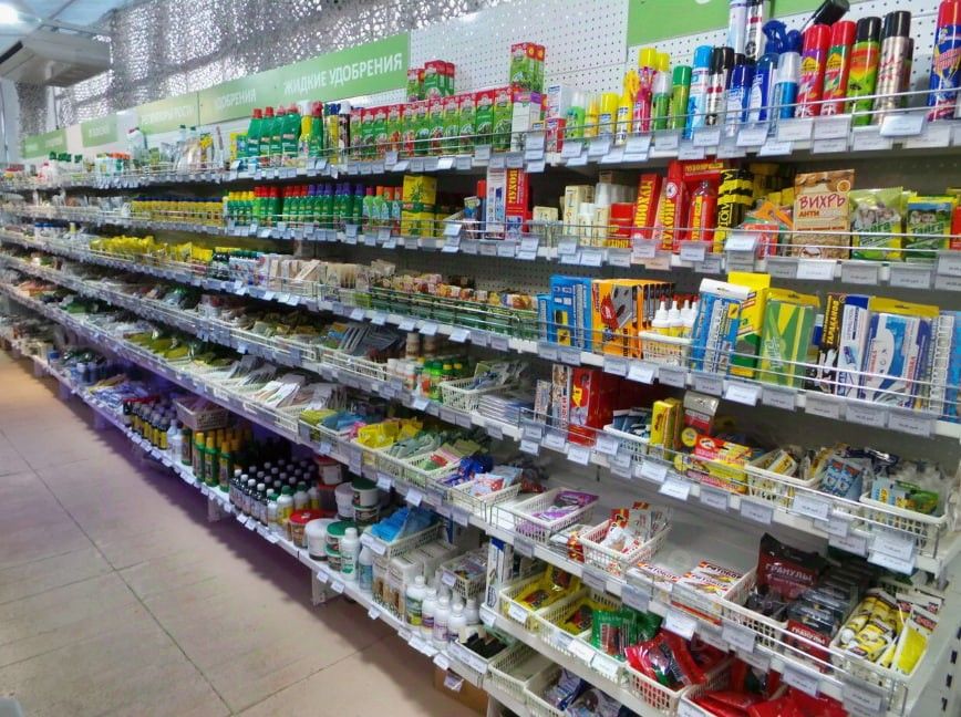 На фото представлена полка в магазине, заполненная разнообразными товарами. Полки организованы в несколько уровней, где размещены упаковки с жидкими удобрениями, средствами для ухода за растениями, а также различными садовыми инструментами. Цветовая палитра включает яркие оттенки упаковок, такие как красный, зеленый, желтый и синий. Общее оформление пространства функциональное, с акцентом на удобство доступа к продуктам. Использованы металлические решетчатые конструкции для хранения товаров, что создает современный и упорядоченный вид.