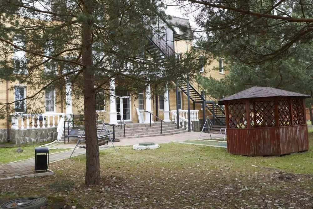 Архитектурный стиль здания выполнен в классическом направлении с кирпичной отделкой. Видны ступени, ведущие к входу, обрамленные белыми колоннами. На заднем плане расположена металлическая лестница, ведущая на второй этаж. Внутренний двор оформлен газоном и мощеной площадкой. На переднем плане выделяется деревянная беседка с решетчатой конструкцией и крышей. Цветовая палитра включает теплые оттенки кирпича и натуральное дерево. Общая атмосфера пространства создает ощущение уюта и комфорта, идеально подходящего для отдыха на свежем воздухе.