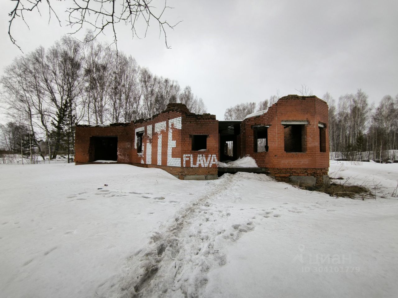 Стены из красного кирпича, частично разрушенные, создают впечатление заброшенного строения. Видны открытые проемы окон и дверей, что придает пространству ощущение незавершенности. Отсутствие крыши и обрушившиеся элементы конструкции усиливают атмосферу запустения. На стенах заметны граффити, в том числе слово 'СДАЕТСЯ', что указывает на возможное назначение здания. Пол окружен снегом, а следы на земле ведут к входу, подчеркивая заброшенность и изоляцию этого места. Цветовая гамма состоит из красного кирпича и белого снега, создавая контраст.