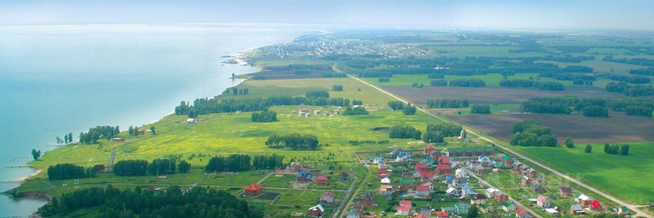 На фото представлен открытый ландшафт с зелеными полями и участками земли. Визуально выделяются небольшие жилые постройки с разнообразными крышами, окрашенными в яркие цвета. Пространство организовано с дорожками, которые соединяют разные участки. Участки земли выглядят ухоженными, с зелеными насаждениями и небольшими садами. Вдалеке виден водоем, который добавляет живописности пейзажу. Архитектурные элементы зданий разнообразны, включая как современные, так и традиционные стили. Цветовая палитра включает зеленые, красные и белые оттенки, создавая гармоничное восприятие пространства.