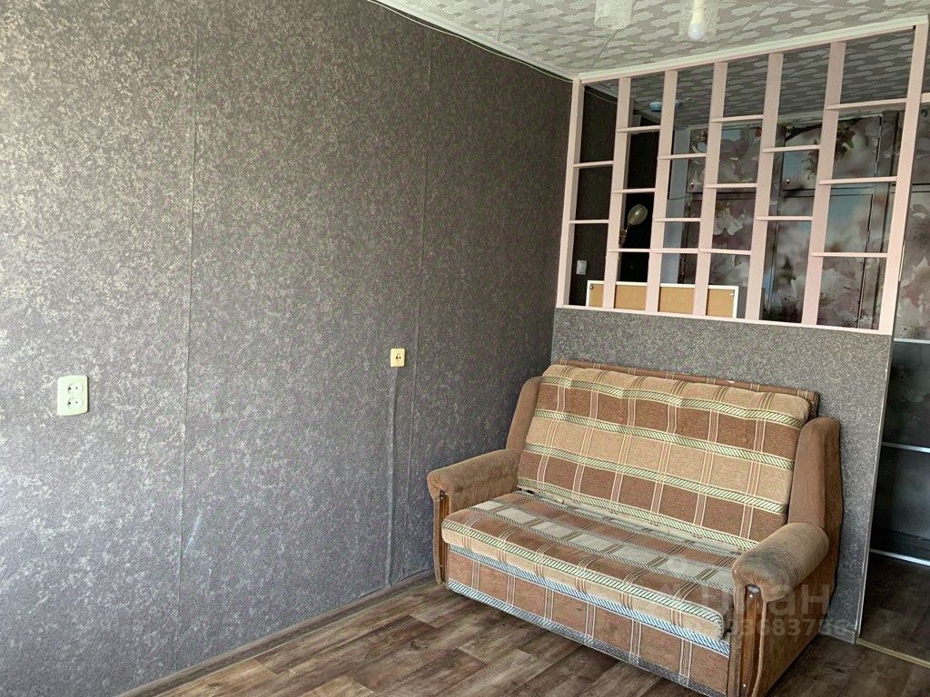 В интерьере виден уголок комнаты с диваном, обитым тканью в коричневых и бежевых тонах. Стены оформлены в сером цвете с текстурированной отделкой, создающей современный вид. На заднем плане расположена перегородка, выполненная из светлого материала, которая разделяет пространство, добавляя легкости и света. Пол покрыт деревянным ламинированным покрытием, что придает уютную атмосферу. Электрические розетки расположены на стенах, обеспечивая удобство для подключения техники. Освещение представлено простыми потолочными светильниками, создающими мягкий свет.