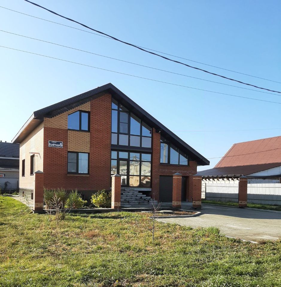 Архитектурный стиль здания выполнен в современном направлении с элементами минимализма. Фасад из кирпича с большими панорамными окнами создает ощущение простора и света. Строение имеет ассиметричную крышу с наклоном, что придает ему уникальный вид. Входная зона оформлена колоннами и лестницей, ведущей к двери. Цветовая палитра включает теплые оттенки кирпича и темные элементы крыши. Основные характеристики включают открытость пространства и гармоничное сочетание материалов, что создает уютную атмосферу. Внешний вид подчеркивает современный подход к жилой архитектуре.