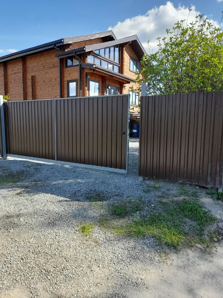 На фотографии представлено загородное строение в современном архитектурном стиле. Деревянный фасад с большими окнами создает ощущение простора и света. Структура имеет два этажа, с наклонной крышей и выступающими элементами. Видно, что входная группа оформлена с использованием стеклянных панелей, что подчеркивает современный дизайн. Огороженная территория со стальными воротами и гравийным покрытием добавляет уюта и приватности. Цветовая гамма теплых древесных оттенков гармонично сочетается с окружающей зеленью, создавая привлекательный внешний вид.